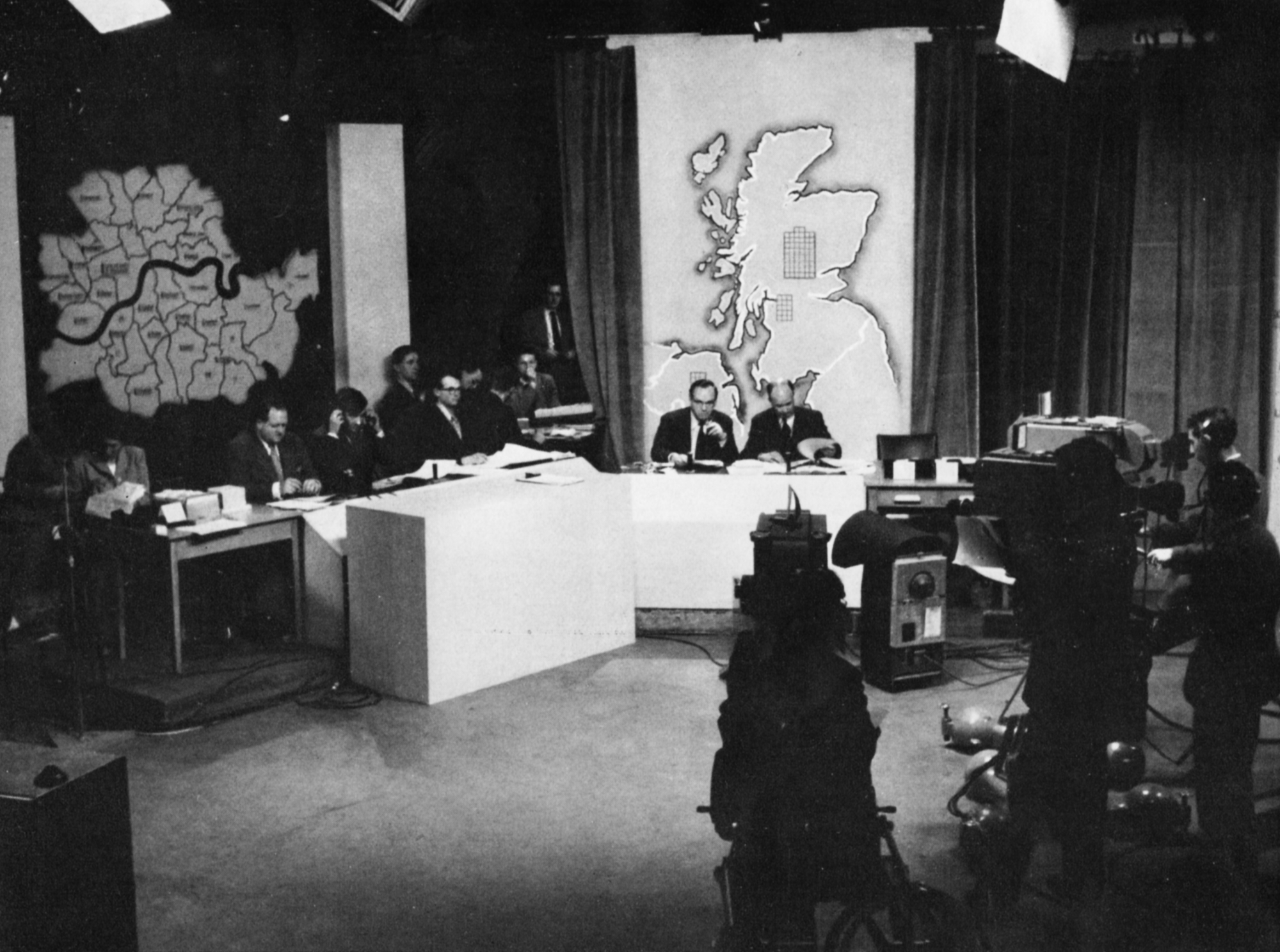 Presenters site behind desks with maps of Scotland and London behind them