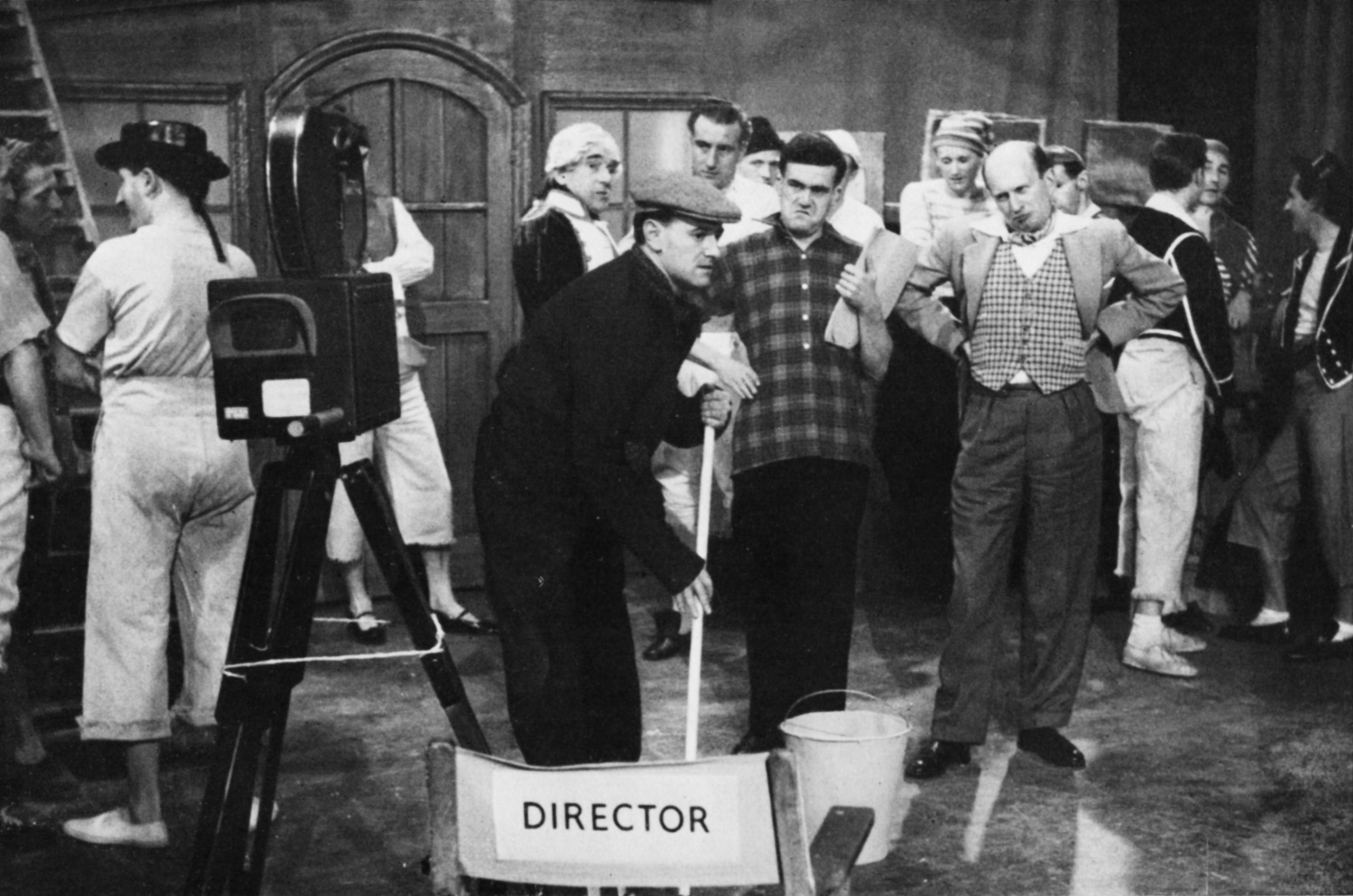 Actor playing actors and stage staff watch a man in a flat cap mop the floor