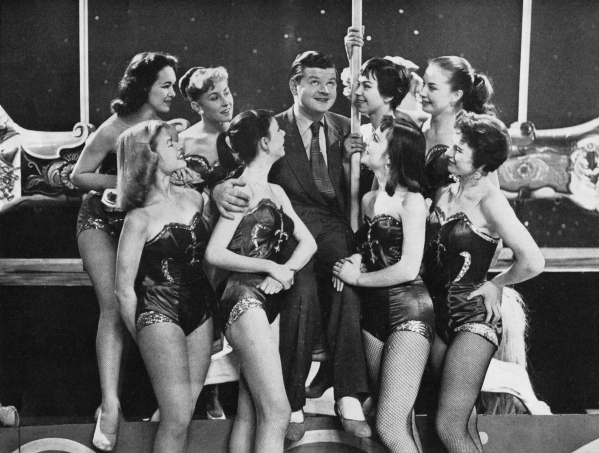 Benny Hill sits on the edge of a carousel whilse surrounded by 8 girls in skimpy costumes