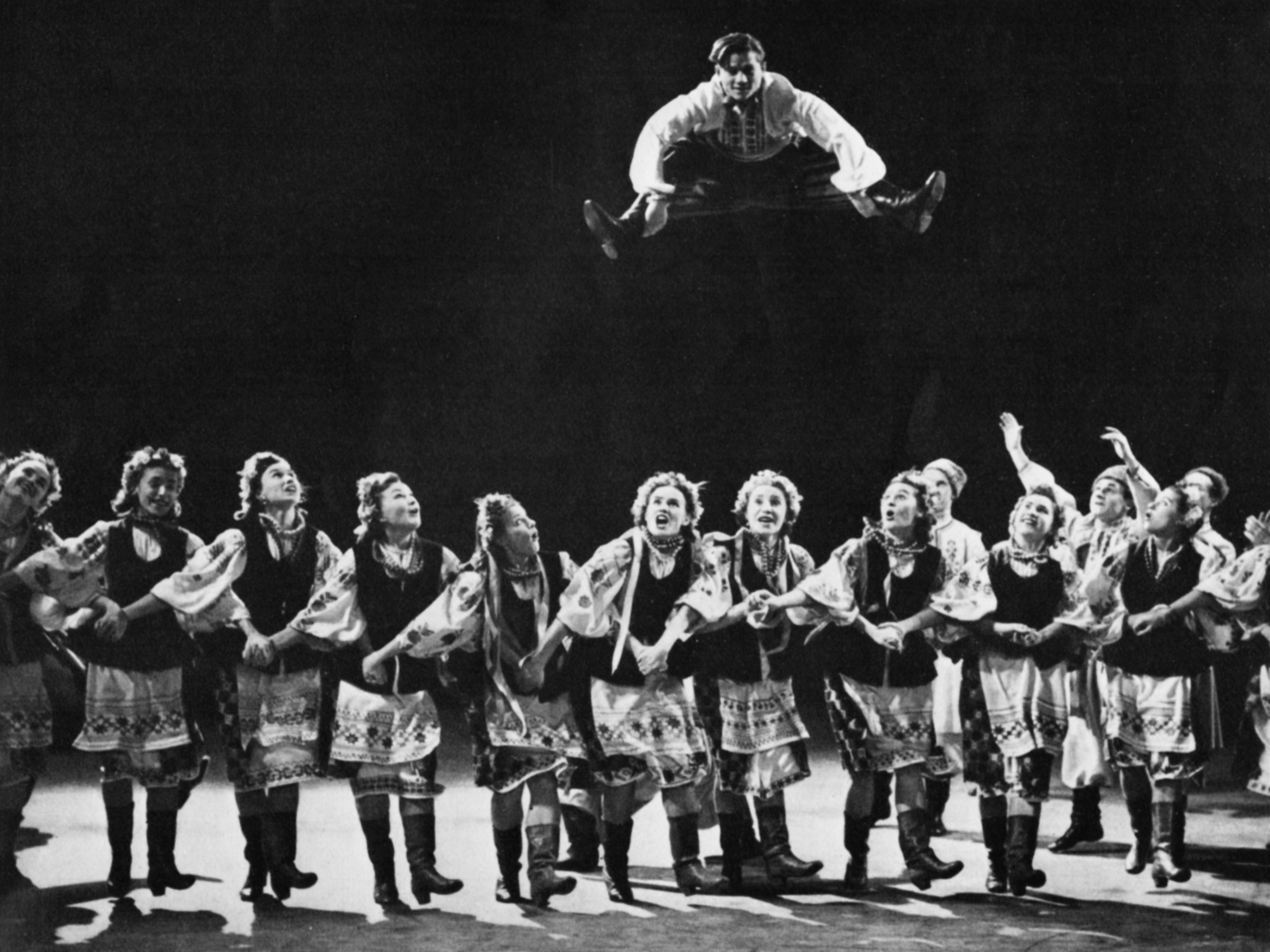 A young male Soviet dancer, his hands on his widely apart legs, soars over 14 other dancers who are holding hands in a line