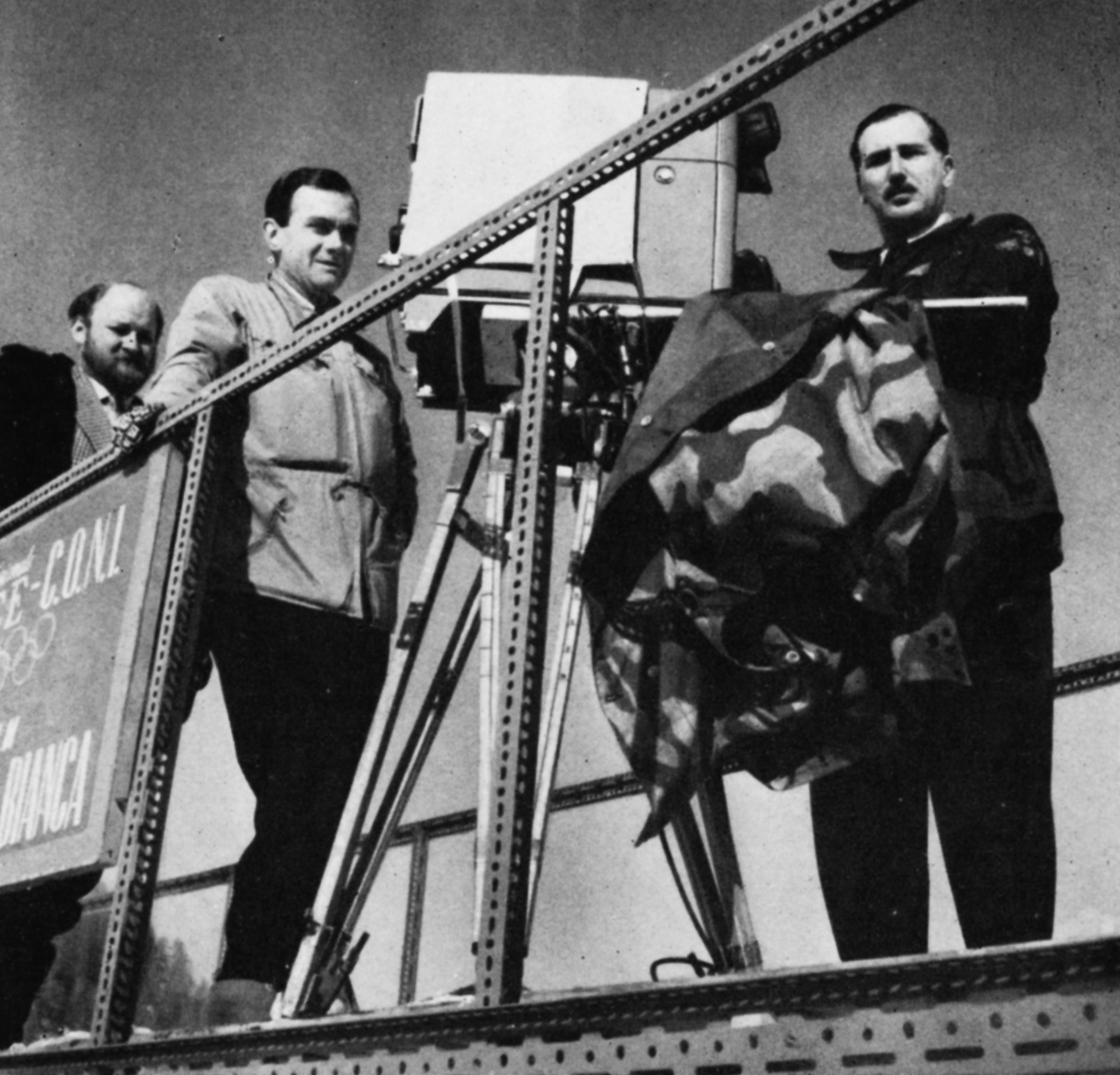 Three men stand beside a camera on an outdoor gantry