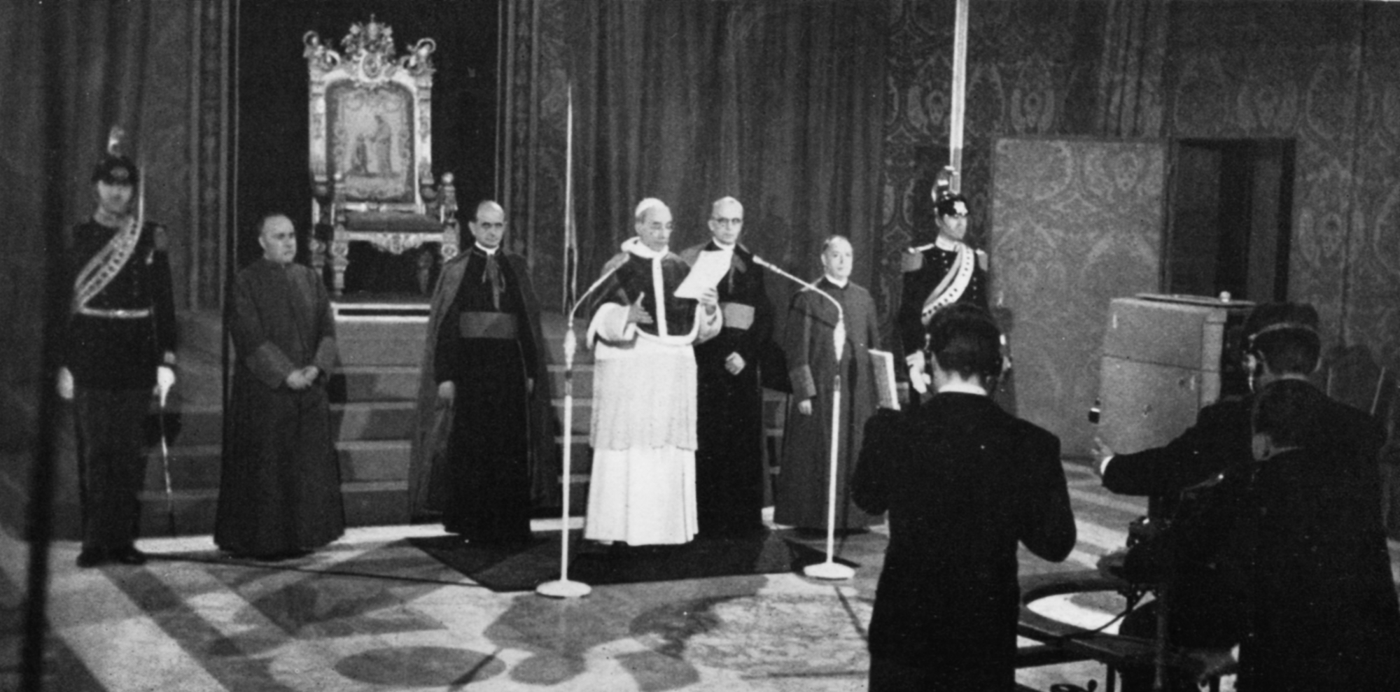 Pope Pius XII reads from a script with two microphones and a camera in front of him