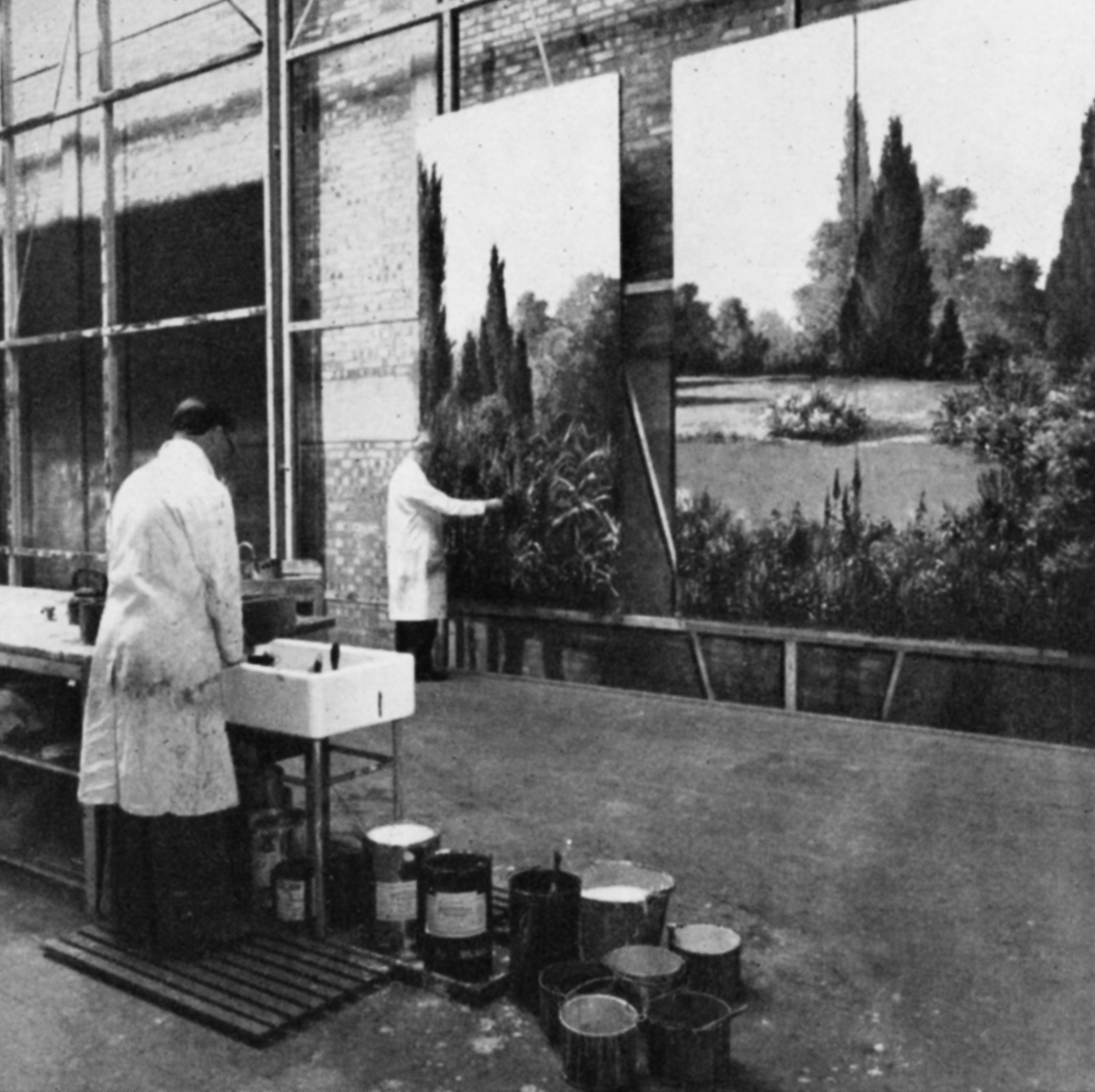 A man paints a garden scene on to a huge background cloth whilst another washes brushes in a sink.