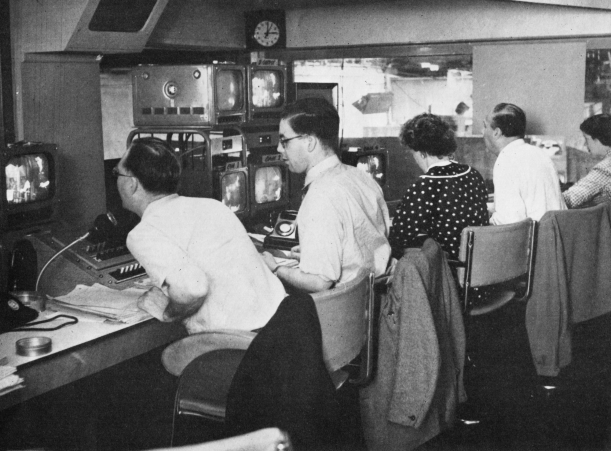 Three men and two women at a control desk covered in monitors.